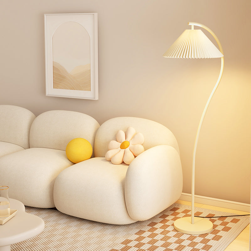 A modern floor lamp with a pleated white shade and curved white stand, glowing warm yellow in a cozy living room setting next to a cream-colored sofa.