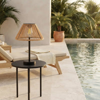 A modern black side table with a table lamp in the foreground, set on a poolside patio with lounge chairs, plants, and a swimming pool in the background.