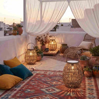A styled outdoor terrace at sunset with white draped curtains, woven rattan lanterns, a wicker lounge chair, patterned rugs, potted plants, and cushions in yellow and teal.