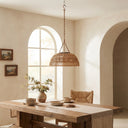A woven natural rattan pendant light hangs over a rustic wooden dining table in a warm, neutral-toned room with arched windows and arches.