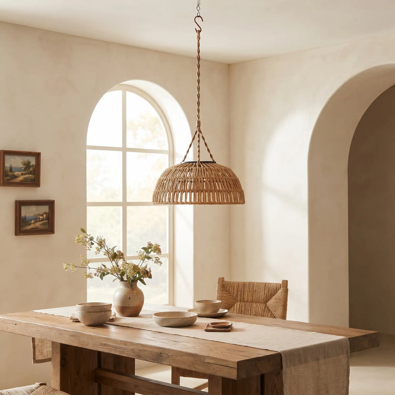A woven natural rattan pendant light hangs over a rustic wooden dining table in a warm, neutral-toned room with arched windows and arches.