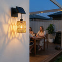 PT0909 solar wall sconce mounted on white stucco wall at dusk, casting warm woven shadow patterns beside wooden dining table where family enjoys wine — cozy alfresco ambiance with IP54 rating and dual solar/USB power.