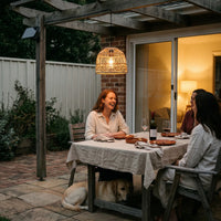 Three women laughing at dinner under PT0913 pendant on brick patio at dusk — dog resting beneath table, illustrating cozy social gatherings enhanced by soft woven light and IP54 weatherproof design.
