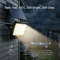 T001ES solar flood light mounted on exterior wall during heavy rain and dark stormy sky, illuminating backyard with trampoline. Bottom icons: IP65 waterproof rating, Breathable Rainproof and Anti-Fog, Iron Housing + Tempered Glass + Iron Bracket. Headline: "Rain. Hail. 45°C. Still Bright. Still Clear."