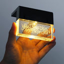 Hand holding a small rectangular light fixture with a black frame and clear glass cover against a gray background.