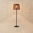 A modern floor lamp with a dark metal stand and a woven wicker shade, glowing with a warm light against a beige wall that casts leaf-like shadows.