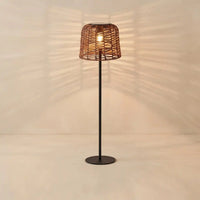 A modern floor lamp with a dark metal stand and a woven wicker shade, glowing with a warm light against a beige wall that casts leaf-like shadows.