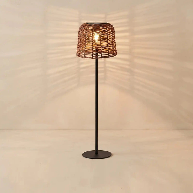 A modern floor lamp with a dark metal stand and a woven wicker shade, glowing with a warm light against a beige wall that casts leaf-like shadows.