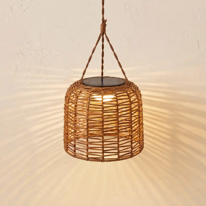 A hanging woven rattan pendant light with a warm glowing bulb inside, suspended by a rope from a ceiling, casting sunburst shadows on a textured beige wall.