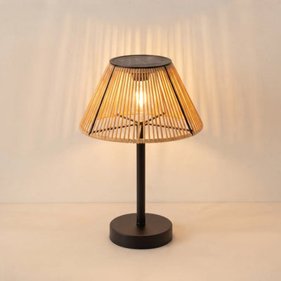 A modern table lamp with a black metal base and a conical woven bamboo shade, illuminated with a warm light that casts soft shadows on the wall behind it.