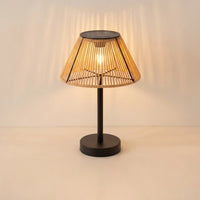 A modern table lamp with a black metal base and a conical woven bamboo shade, illuminated with a warm light that casts soft shadows on the wall behind it.