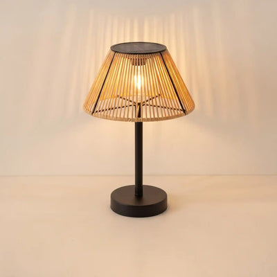 A modern table lamp with a black metal base and a conical woven bamboo shade, illuminated with a warm light that casts soft shadows on the wall behind it.