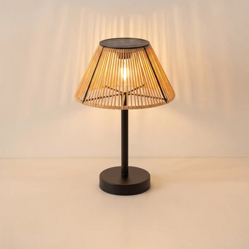 A modern table lamp with a black metal base and a conical woven bamboo shade, illuminated with a warm light that casts soft shadows on the wall behind it.