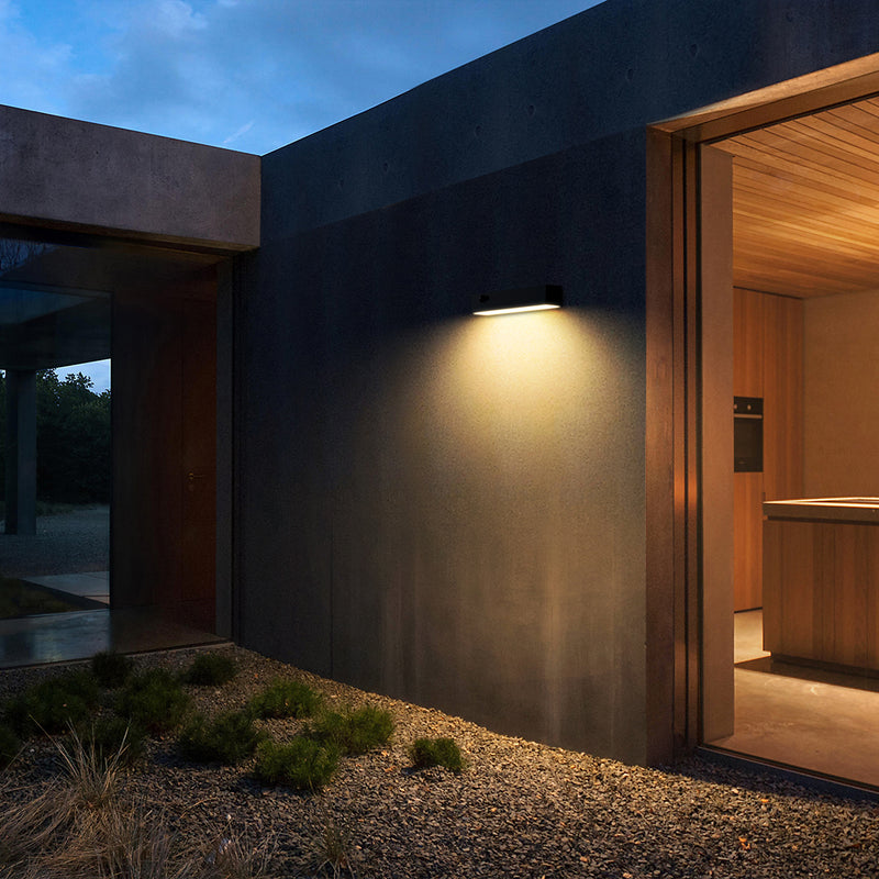 Wall-mounted outdoor light fixtures illuminating a concrete building facade at dusk, with a wooden door, deck, grass, and sky in the background.