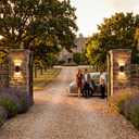 Couple standing next to a vintage car at the entrance of a grand estate with stone pillars and lavender bushes.