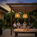 Two woven pendant lights hanging under a wooden pergola on a patio, illuminating a family sitting underneath in a lush backyard setting.