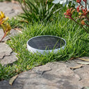 Close-up of a solar ground light installed in green grass next to stone pavers, featuring a sleek white round design with a black solar panel top, blending into the garden landscape.