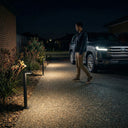 Man walking gravel driveway at night beside Toyota Land Cruiser, Outway PT0724 Nivra aluminium solar bollard lights casting warm amber pools along garden border