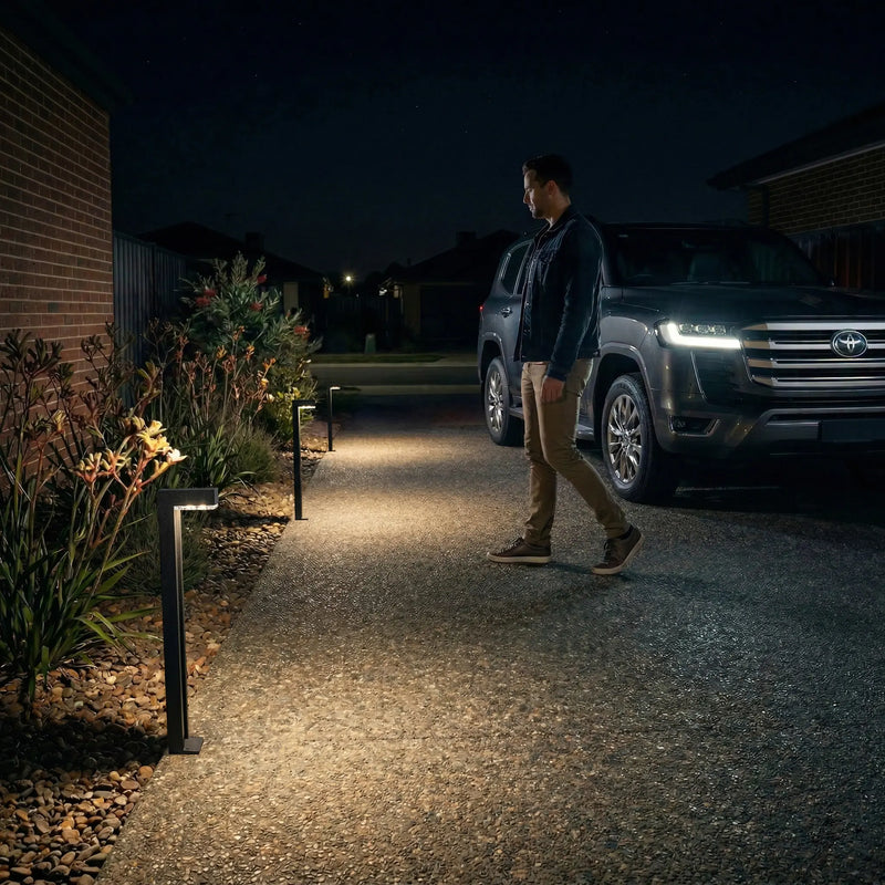 Man walking gravel driveway at night beside Toyota Land Cruiser, Outway PT0724 Nivra aluminium solar bollard lights casting warm amber pools along garden border