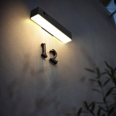 A modern rectangular wall-mounted light bar with a glowing warm white LED, mounted on a textured gray wall above the house number 12.