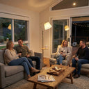 FL068 gold floor lamp with wood tray as the centrepiece of a warm living room gathering, amber light casting a soft glow over two couples sharing wine and a cheese board