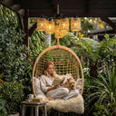 Woman reading a book with a dog on her lap in a hanging chair surrounded by plants.