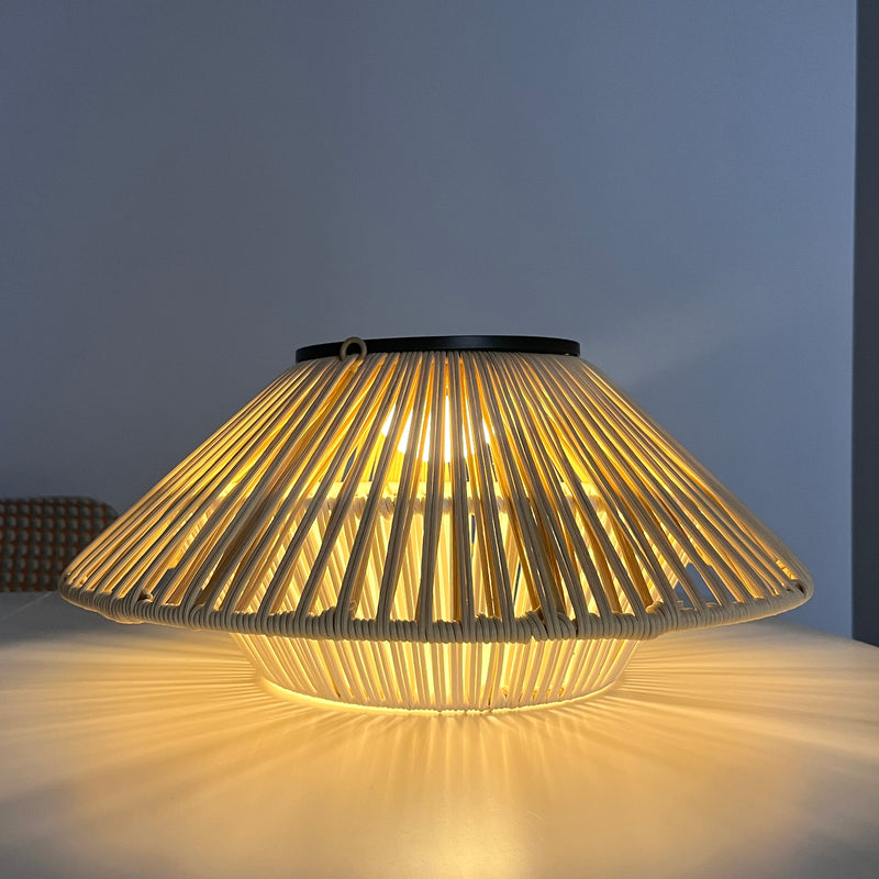 A modern table lamp with a wide, conical shade made of vertical woven slats, glowing with a warm yellow light against a dark blue-gray background.