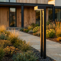 Outway PT0724 Nivra solar bollard light standing in mixed garden border at dusk, black aluminium square column glowing warm gold, modern house backdrop