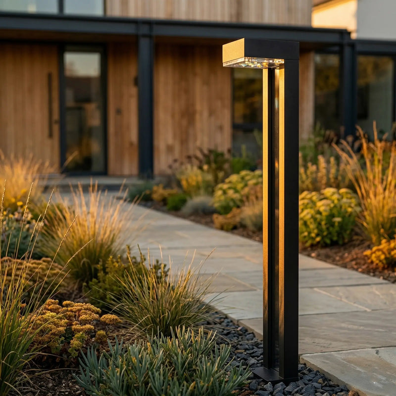 Outway PT0724 Nivra solar bollard light standing in mixed garden border at dusk, black aluminium square column glowing warm gold, modern house backdrop