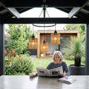 Woman reading a newspaper in a modern outdoor setting with plants and a house in the background.