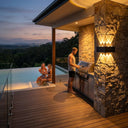 An outdoor kitchen scene at dusk featuring a man grilling by a stone pillar illuminated by the Grivo solar wall light, while two women relax in an infinity pool overlooking a forest and city skyline in the distance.