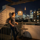 Man relaxing on a rooftop balcony at night with PT0907 Solar Pendant Light glowing overhead, casting woven shadows on the wall. Sydney Tower visible in background. Warm 3000K ambiance perfect for urban outdoor living spaces.