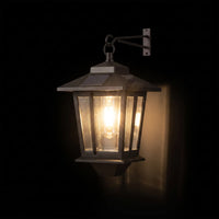 A solar-powered outdoor wall lantern light with a dark metal frame and glass panels, glowing with a warm light against a dark background.