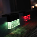 A close-up of a rectangular solar pathway light with a black housing and a glowing green illuminated panel, mounted on a dark surface next to a textured mat. A second red light is visible in the background.