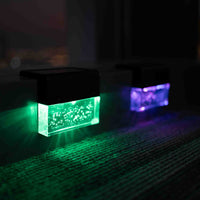 Two rectangular solar-powered light projectors casting green and purple light beams onto a dark carpeted floor at night.