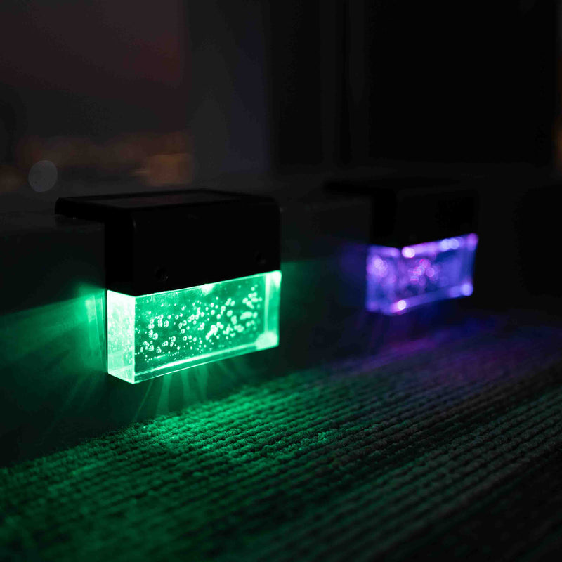 Two rectangular solar-powered light projectors casting green and purple light beams onto a dark carpeted floor at night.