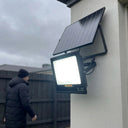 Close-up of T001ES 300W solar flood light mounted on white render wall in overcast winter conditions, man in puffer jacket walking past in background, light glowing bright white. Shows 270° bracket and separate solar panel mount.