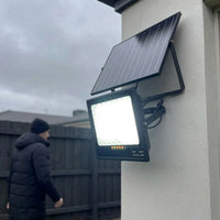 Close-up of T001ES 300W solar flood light mounted on white render wall in overcast winter conditions, man in puffer jacket walking past in background, light glowing bright white. Shows 270° bracket and separate solar panel mount.