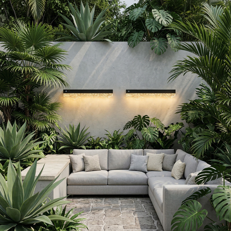 Solar Wall Lights glowing warm amber 3000K on grey rendered feature wall, set in lush tropical outdoor living space with L-shaped sectional sofa, monstera and palm plants, stone paver patio