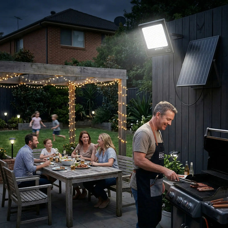 Australian family BBQ night scene: man grilling sausages while family and friends laugh at outdoor table with fairy lights, T001ES solar flood light mounted on fence providing bright overhead illumination. Kids playing in background on lawn.