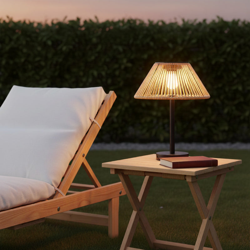 A lit table lamp with a warm glowing bulb and a pleated beige shade sits on a small wooden side table next to a lounge chair in an outdoor garden setting at dusk.