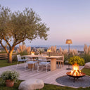 Outdoor patio with floor lamp next to a wooden table, chairs, and fire pit by a tree at dusk.