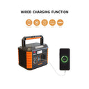 A portable power station with orange and black ribbed sides, shown wired to charge a smartphone. Above the product, the text reads 'WIRED CHARGING FUNCTION' with orange icons indicating compatible devices: phones, cameras, GPS devices, laptops, tablets, and cars.