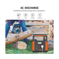 An outdoor product demonstration showing a portable power station branded URANI on the ground next to a person using a chainsaw to cut a log. The power station is orange and black with a built-in handle and multiple output ports, including a power cord plugged in.