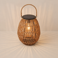 A large woven rattan lantern with a handle sits on terracotta tiles in a white arched courtyard, casting warm radial shadows near an olive tree and built-in bench.