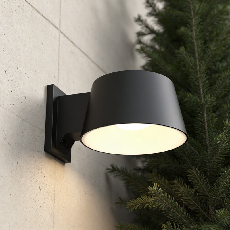 A matte black cylindrical wall-mounted outdoor light fixture casting a warm glow, shown mounted on a light concrete wall next to a green pine tree.