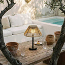 A modern table lamp with a warm glowing bamboo slatted shade and a black base, styled on a rustic wooden table in an outdoor patio setting with a white sofa, a small pool, and terracotta pots.