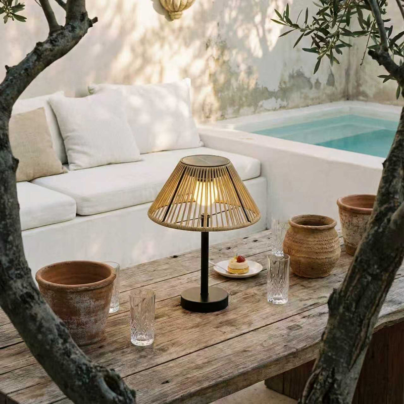 A modern table lamp with a warm glowing bamboo slatted shade and a black base, styled on a rustic wooden table in an outdoor patio setting with a white sofa, a small pool, and terracotta pots.