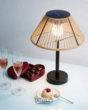 A modern table lamp with a natural rattan shade and a black base, featuring a dark blue solar panel on the top of the shade. The lamp is styled on a light blue table next to two champagne flutes, a heart-shaped box of chocolates, and a small dessert plate, with the 'OutWry' brand logo at the bottom.