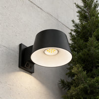 A modern black outdoor wall sconce with a cylindrical shade, mounted on a textured concrete wall next to a green evergreen tree. The light is turned on, casting a warm glow downward.